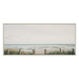 Framed canvas showing a pale sky over a calm ocean with a sandy beach path bordered by wooden posts and rope fencing through tall green grass.