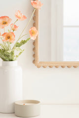 Partial view of a Beadbrook arched wall mirror with rounded bead detailing on a light wood frame, next to a white vase with peach poppy flowers and a small beige candle holder.