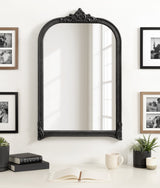 Arched wall mirror with an ornate black frame featuring elegant scrollwork at top and bottom, reflecting a window's light in a simple white room.