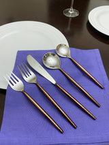 Set of hammered stainless steel flatware with brown hammered handles, including two forks, one knife, and two spoons, arranged on a purple napkin next to white plates.