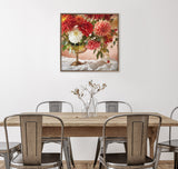 Square framed canvas of a floral arrangement with large red, white, and pink flowers in a gold vase on a white cloth, hanging above a wooden table with metal chairs.