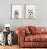Two framed animal portraits on a light gray brick wall above a brown leather sofa; one features a fluffy owl, the other a gray wolf, both on white backgrounds.