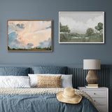 Two framed landscape paintings above a bed with blue bedding; left painting shows soft clouds in pastel tones, right painting depicts green trees and hills under a cloudy sky.