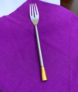 Silver fork with a slender handle featuring a gold-toned end, resting on a purple cloth surface with a white plate edge visible above.