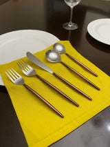 Five-piece hammered stainless steel flatware set with brown handles arranged on a bright yellow cloth over a dark wood table, surrounded by white plates and a wine glass.