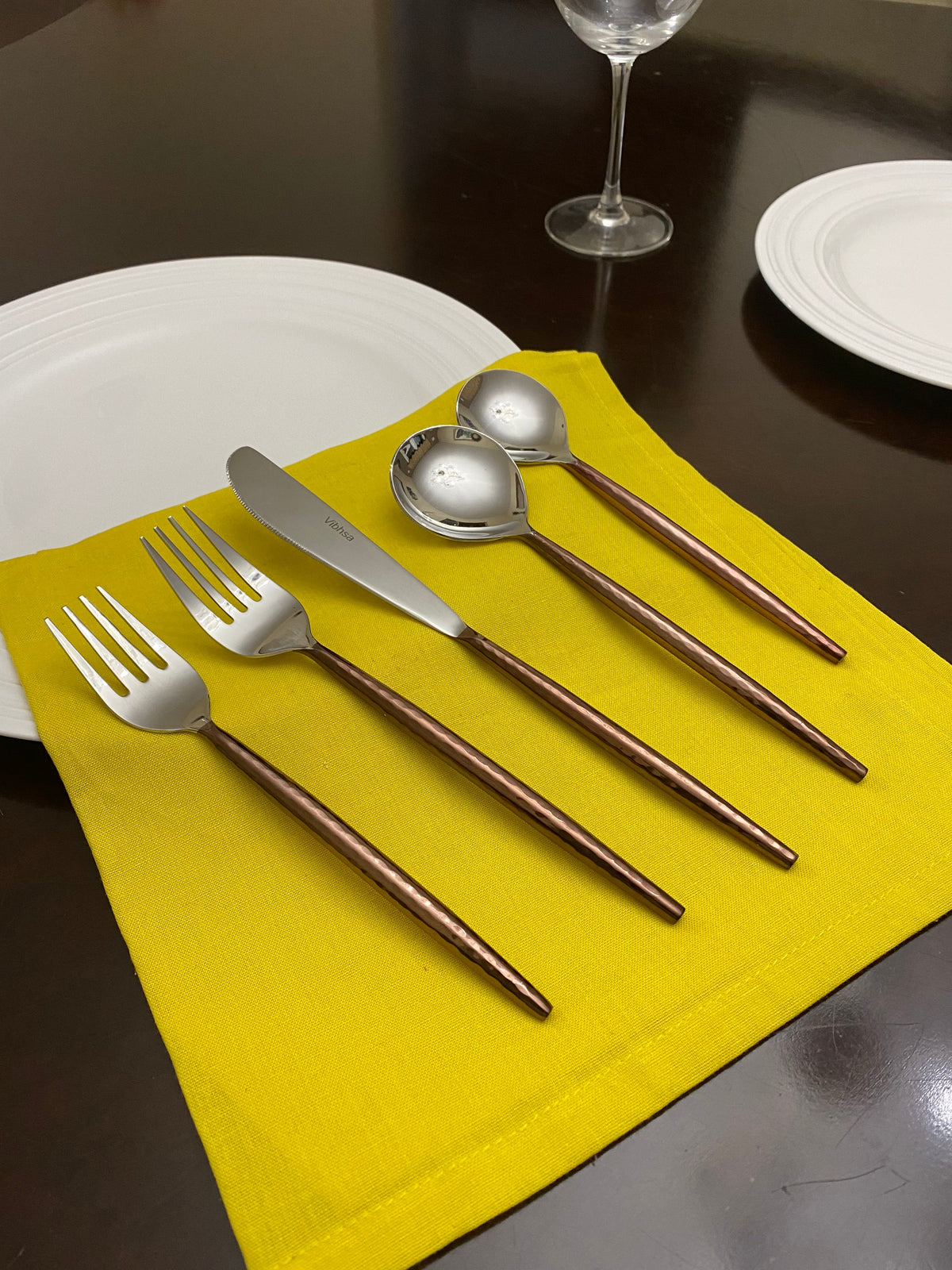 Five-piece hammered stainless steel flatware set with brown handles arranged on a bright yellow cloth over a dark wood table, surrounded by white plates and a wine glass.