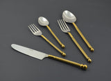 Set of stainless steel flatware with twisted golden handles including a knife, two forks, and two spoons displayed on a dark background.