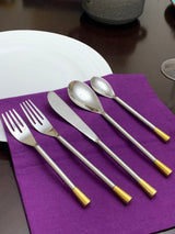 Five-piece silver flatware set with sleek handles featuring gold-colored ends, arranged on a purple cloth next to a white plate on a dark wooden table.