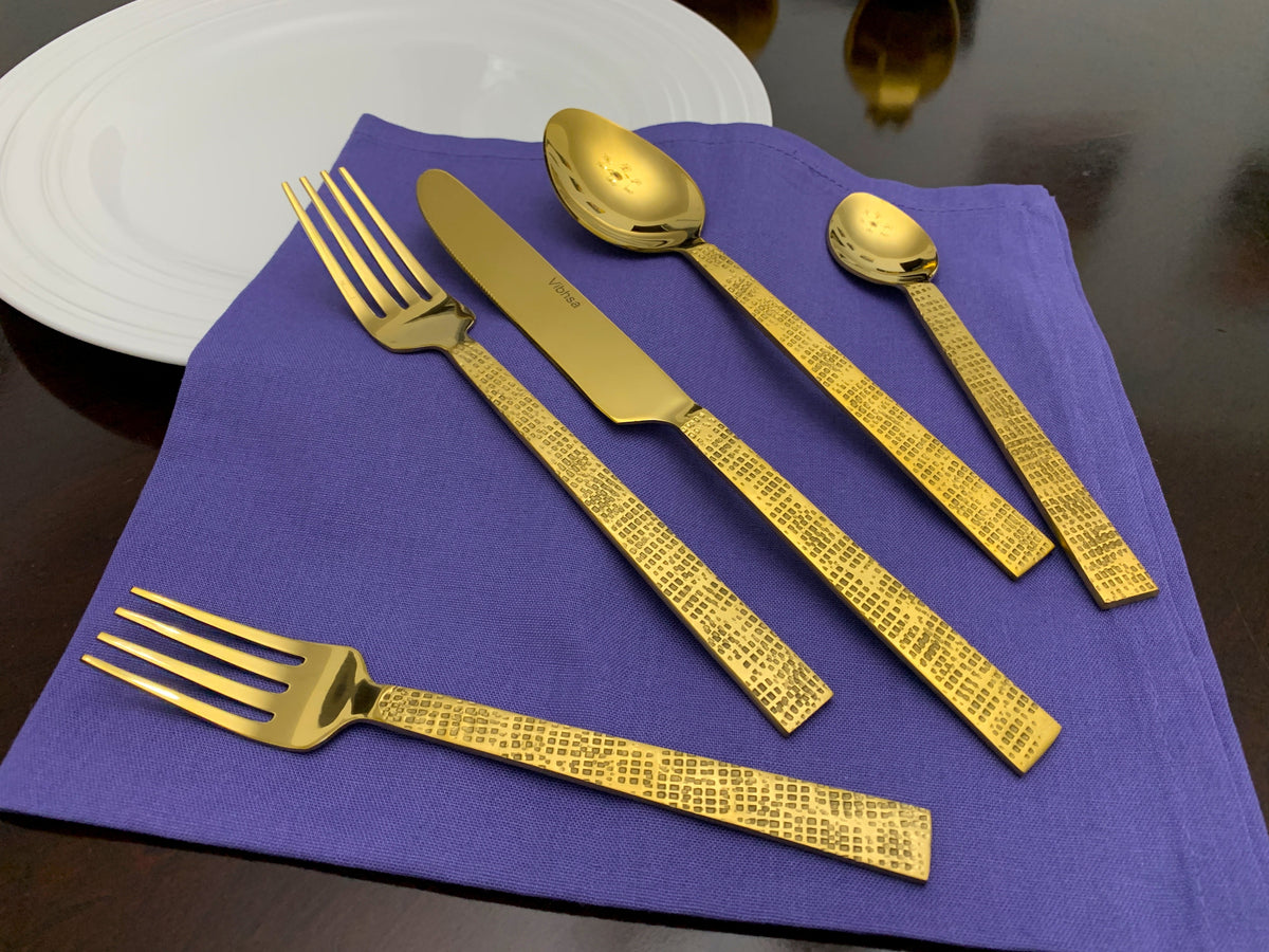 Five-piece gold flatware set with textured Egyptian design handles resting on a purple cloth beside a white plate on a dark wooden table.