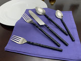Five-piece stainless steel flatware with black twisted swirl handles arranged on a purple cloth near white plates on a dark wooden surface.