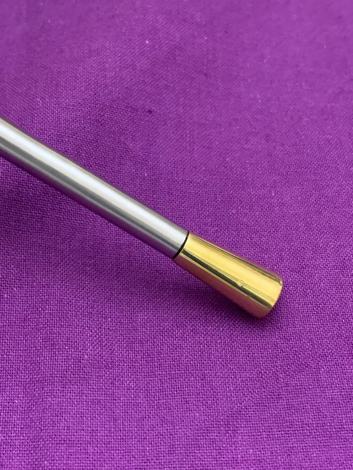 Close-up of a silver metal rod with a polished gold-colored tapered tip, resting on a textured purple fabric background.