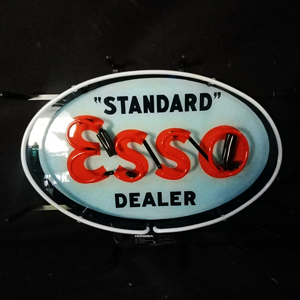 Oval blue neon sign with white border reading "STANDARD" in black above large red neon letters spelling "ESSO" and "DEALER" in black below, shown against a black background.