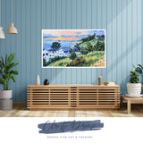 Colorful landscape painting of coastal houses on green hills under a blue sky with pink clouds, displayed above wood slat cabinet against blue paneled wall.