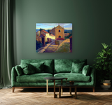 Canvas print of a sunlit Portuguese street with an ochre church, white houses, and blue sky, hanging on a muted green wall above a dark green velvet sofa.