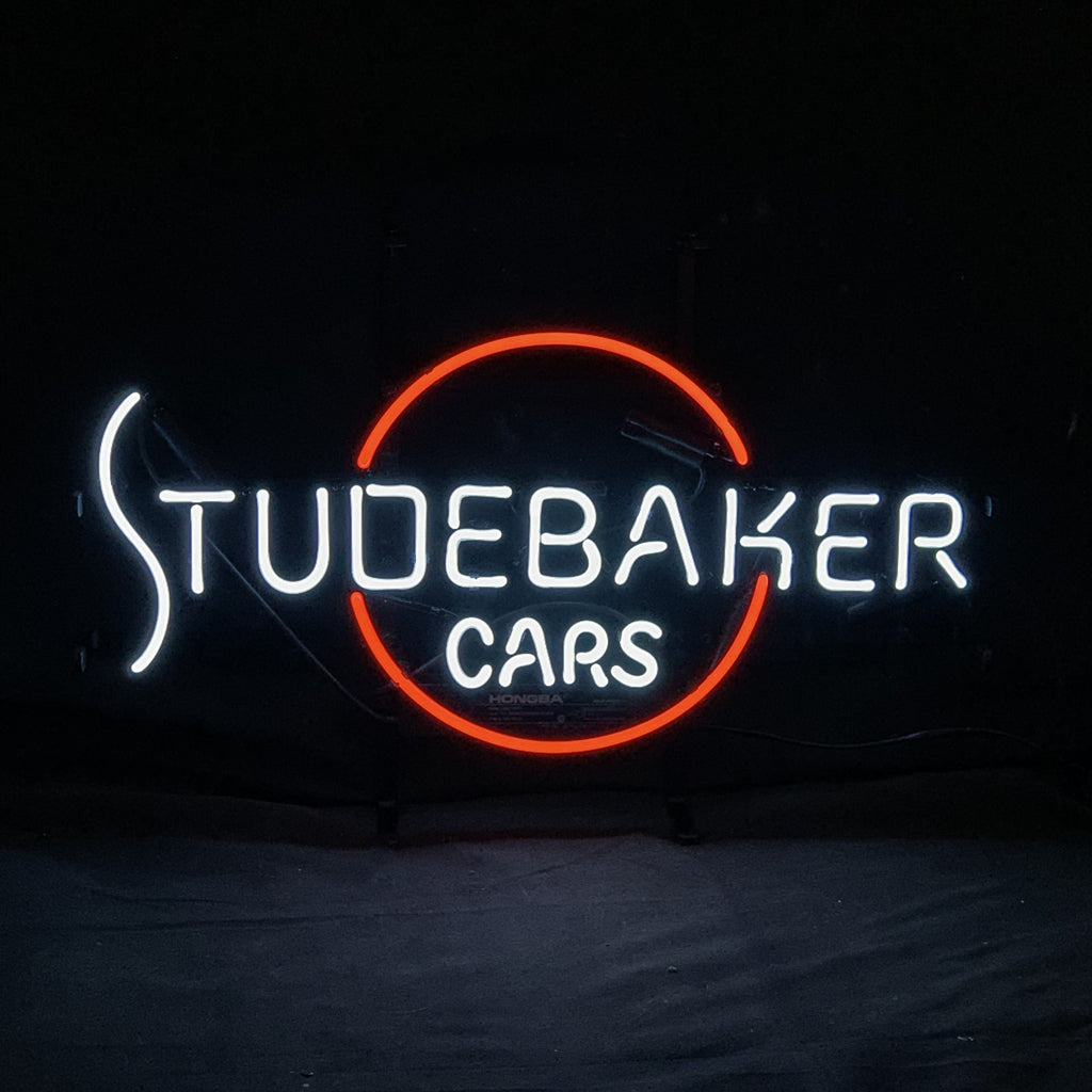 Neon sign glowing with white text 'STUDEBAKER CARS' and an incomplete red circle behind the word 'BAKER' on a black background.