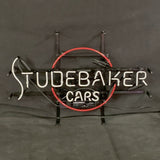 Neon sign with white letters spelling 'STUDEBAKER' above smaller 'CARS' in white, encircled by a red neon ring on black background.