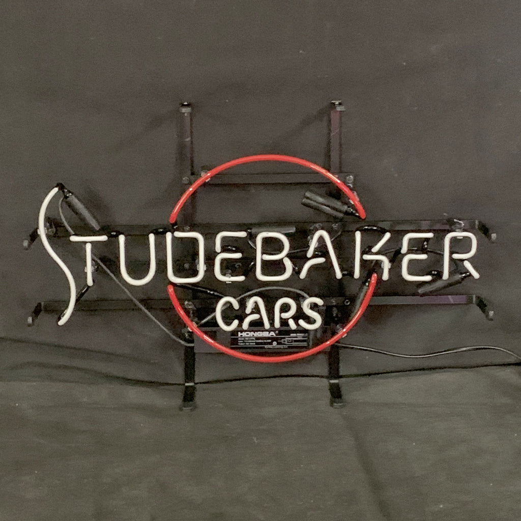 Neon sign with white letters spelling 'STUDEBAKER' above smaller 'CARS' in white, encircled by a red neon ring on black background.