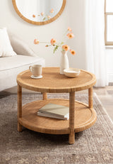 Round rattan coffee table with woven texture, featuring a lower shelf holding books, styled with a white vase of peach flowers, cream bowl, and mug on top
