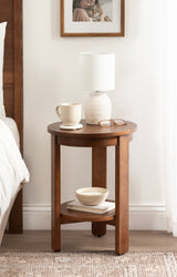 Round wooden side table with warm brown finish, holding a textured white lamp, a pale beige mug, and reading glasses, plus a lower shelf with a bowl and books.