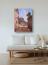 Impressionist-style painting of a sunlit path flanked by warm-toned buildings and lush greenery, mounted on a white wall above a minimalist beige sofa.