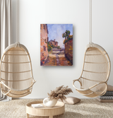 Painting of a sunlit narrow path flanked by rustic buildings and stone walls under a bright blue sky, hung on a white wall between two wicker hanging chairs.