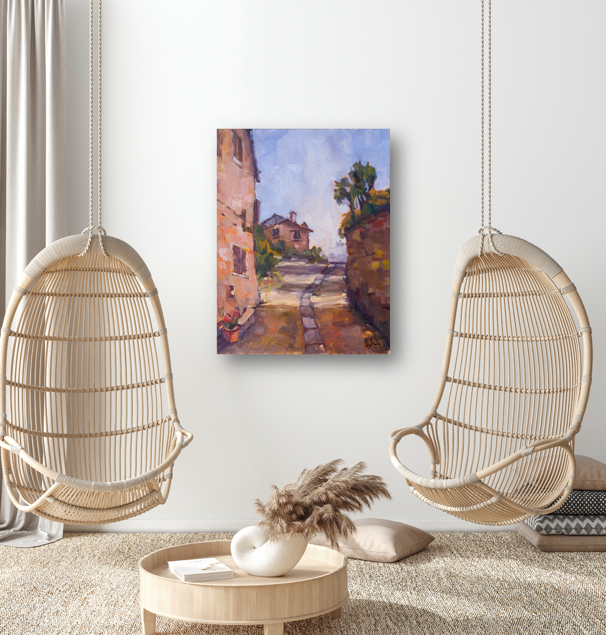 Painting of a sunlit narrow path flanked by rustic buildings and stone walls under a bright blue sky, hung on a white wall between two wicker hanging chairs.