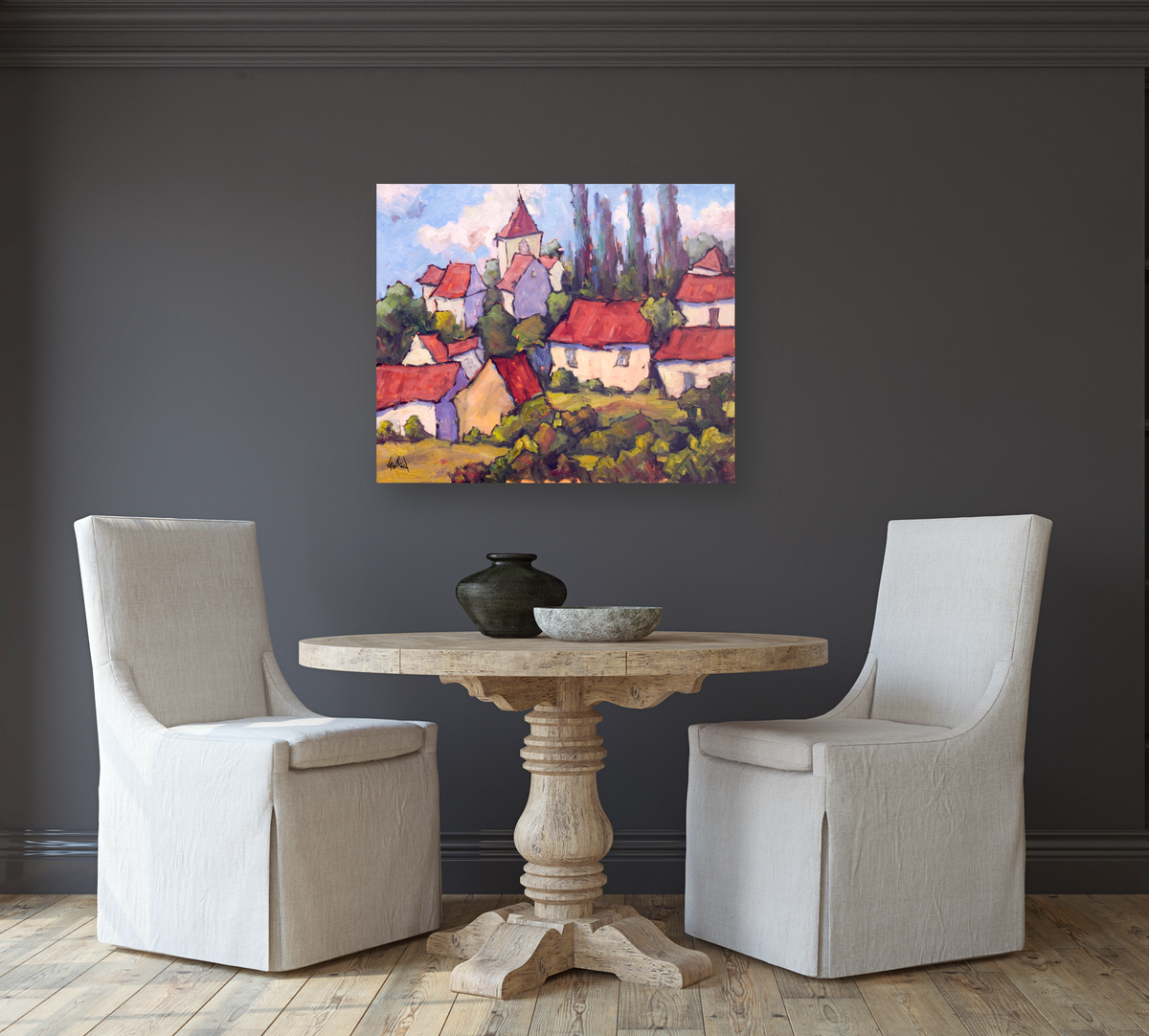 Colorful impressionist painting of red-roofed houses and green trees, hung on a dark gray wall above a round wooden table with two beige upholstered chairs.