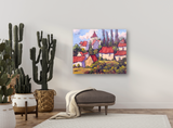 Canvas print of a colorful village scene with red rooftops and green trees, hung on a light wall above a dark cushioned bench with a beige throw.