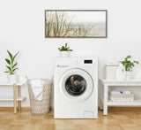 Minimalist laundry room with a white front-loading washer centered between a wicker basket and a white table, featuring a framed beach grass print above the appliance.