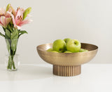 Brushed gold-toned ribbed metal bowl holding five green apples, placed on a white surface next to a clear vase with pink lilies and green stems.