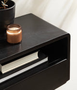 Black rectangular side table with rounded edges and an open storage shelf holding two books, topped with a brown candle and a black plant pot.