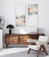 Two vertical abstract landscape paintings with soft beige, blue, and brown hues in thin white frames above a mid-century modern wood sideboard, beside a modern chair and decor.