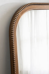 Curved corner of an arched wall mirror with a wood frame featuring a beaded decorative edge in warm brown tones, reflecting sheer white curtains.
