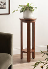 Tall round wood side table with a warm brown finish, featuring three straight legs connected by a circular lower shelf, holding a white pot with green leafy plant.