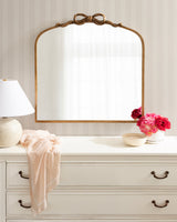 Arched mirror with a golden ribbon-shaped top frame mounted above a white dresser with dark handles, decorated with a white lamp, soft pink fabric, and pink flowers in a vase.