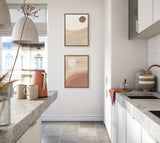 Two framed canvas artworks with sandy and caramel tones, featuring abstract lore moon shapes, hung vertically on a white kitchen wall between beige and gray countertops.