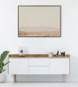 Framed muted desert landscape painting depicting distant mountains and sparse vegetation, hanging above a minimalist wooden sideboard with white drawers and small decor pieces.