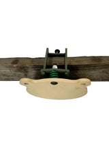 Front view of a shooting target mounted on a wooden beam, featuring a light-colored shaped plate attached with a green metal bracket and spring mechanism.