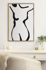 Black and white abstract line drawing of a female torso in a slim wooden frame hanging on a white wall above a white desk with decorative items and a plant.