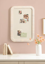 Rectangular fabric pinboard with rounded white frame on a pale pink wall, displaying four photos and a green envelope, above a white surface with books and a vase of light orange flowers.