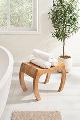 Curved wooden stool with smooth finish holding three rolled white towels, positioned on light tile floor near a potted green leafy plant and window.