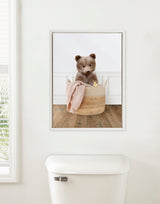Framed canvas showing a fuzzy bear cub sitting inside a woven laundry basket with a pink blanket on wooden floor against a white wall with panel molding.