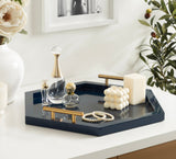 Glossy navy blue hexagonal tray with gold handles holding decorative glass bottles, a pearl necklace, and two textured white candles on a white countertop