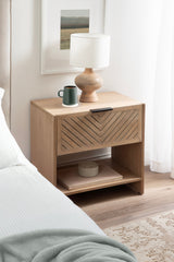 Light wood bedside table with a chevron-patterned drawer and bottom shelf holding books and a white round container, topped with a wooden lamp and green mug.