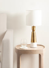 Modern table lamp with white cylindrical shade and tapered brass and white base on a light wood side table beside a beige sofa, with a glass of water on a coaster.