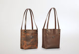 Two leather tote bags with long straps, one in distressed brown with a front patch pocket and logo, the other in smooth dark brown with similar features, displayed upright
