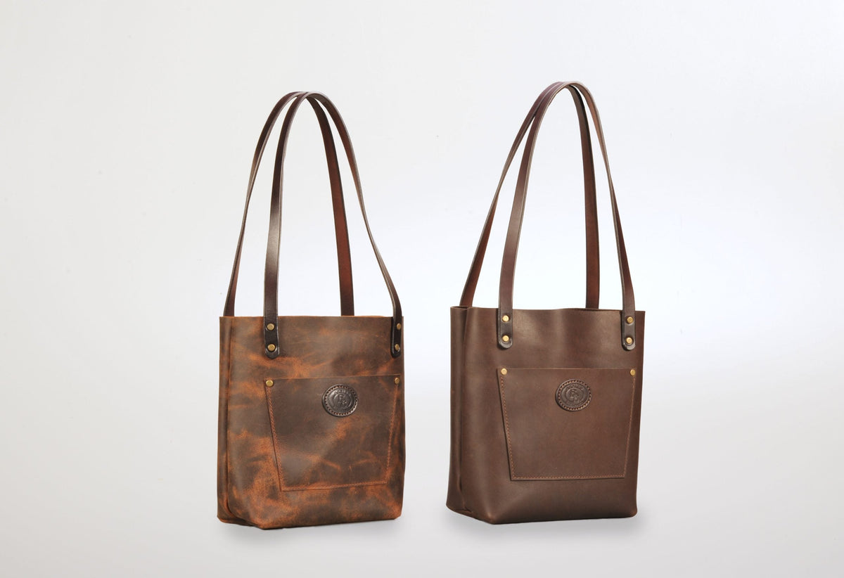Two leather tote bags with long straps, one in distressed brown with a front patch pocket and logo, the other in smooth dark brown with similar features, displayed upright