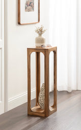 Tall, narrow wooden side table with a hollow central space displaying a decorative vase, topped with a beige ceramic vase holding dried flowers and sunglasses.