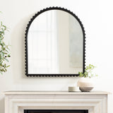 Arched wall mirror with a black scalloped frame mounted above a white marble mantel decorated with a round white textured vase and a small beige candle.