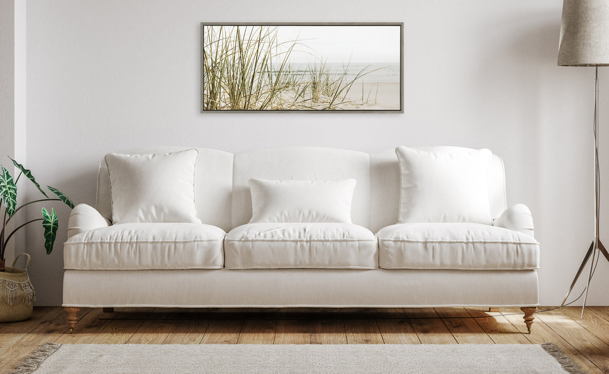 White cushioned sofa with three pillows below a rectangular framed canvas featuring tall beach grass and a muted sandy beach background, in a softly lit living room.